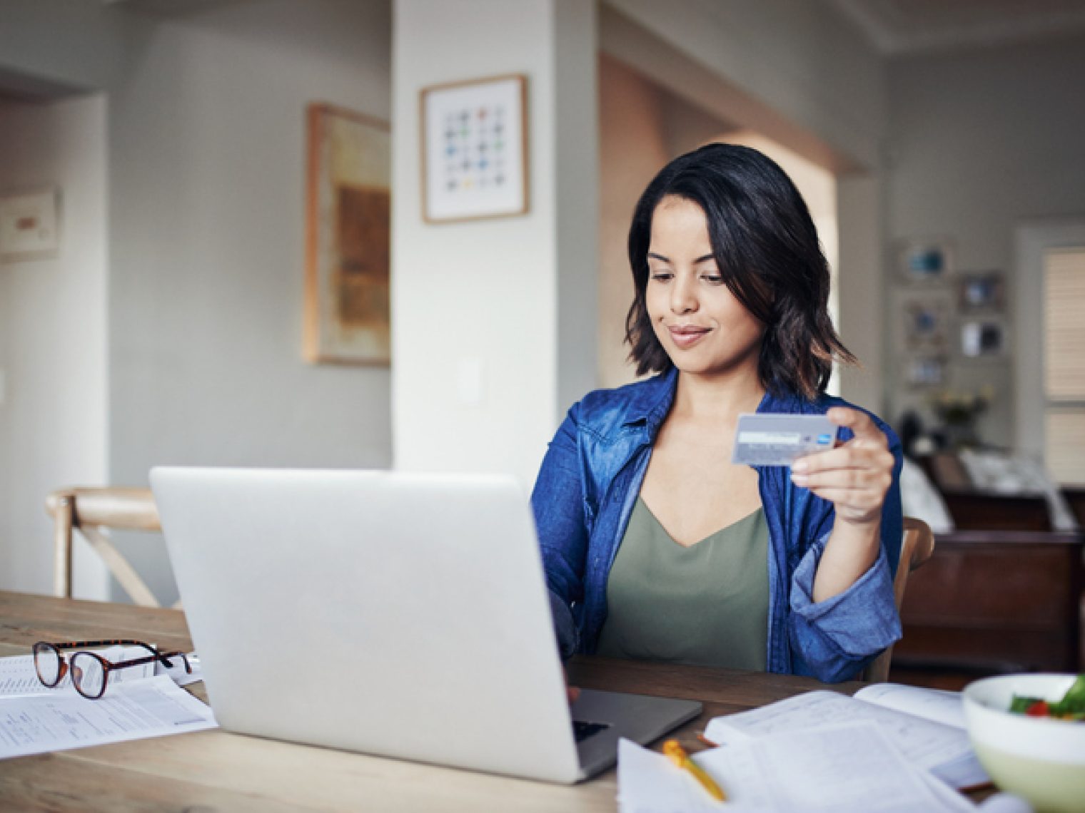 A smiling woman holding a credit card looks at her laptop, surrounded by papers and a pair of glasses on a table in a well-lit home interior.