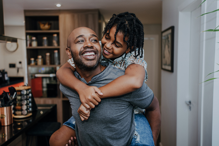 A joyful moment as a father and daughter share a loving embrace, with the child riding piggyback, both smiling in a cozy home setting.