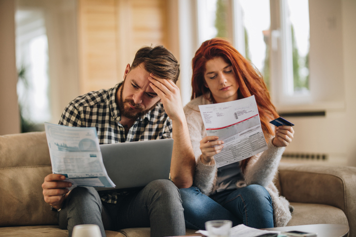 A worried couple is looking at their finances with a sense of concern. The man is holding a document and touching his forehead while the woman is holding another document and a credit card.