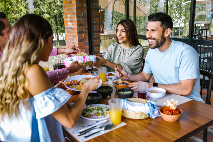 A group of friends enjoys a meal together outdoors, sharing food and smiles at a table filled with various dishes and drinks.