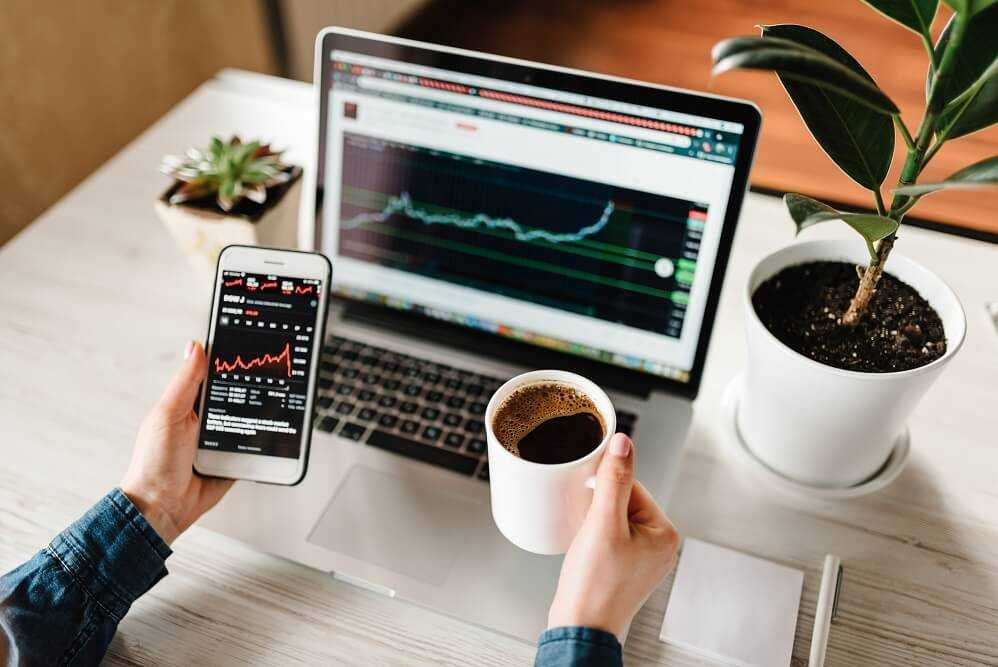 Analyzing stocks on a computer and cell phone with a coffee cup.