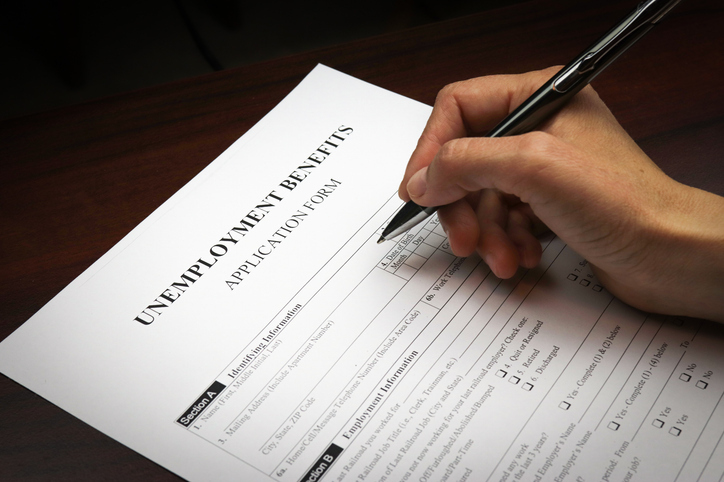 A person's hand is filling out an "Unemployment Benefits Application Form," indicating they are applying for government assistance due to unemployment.